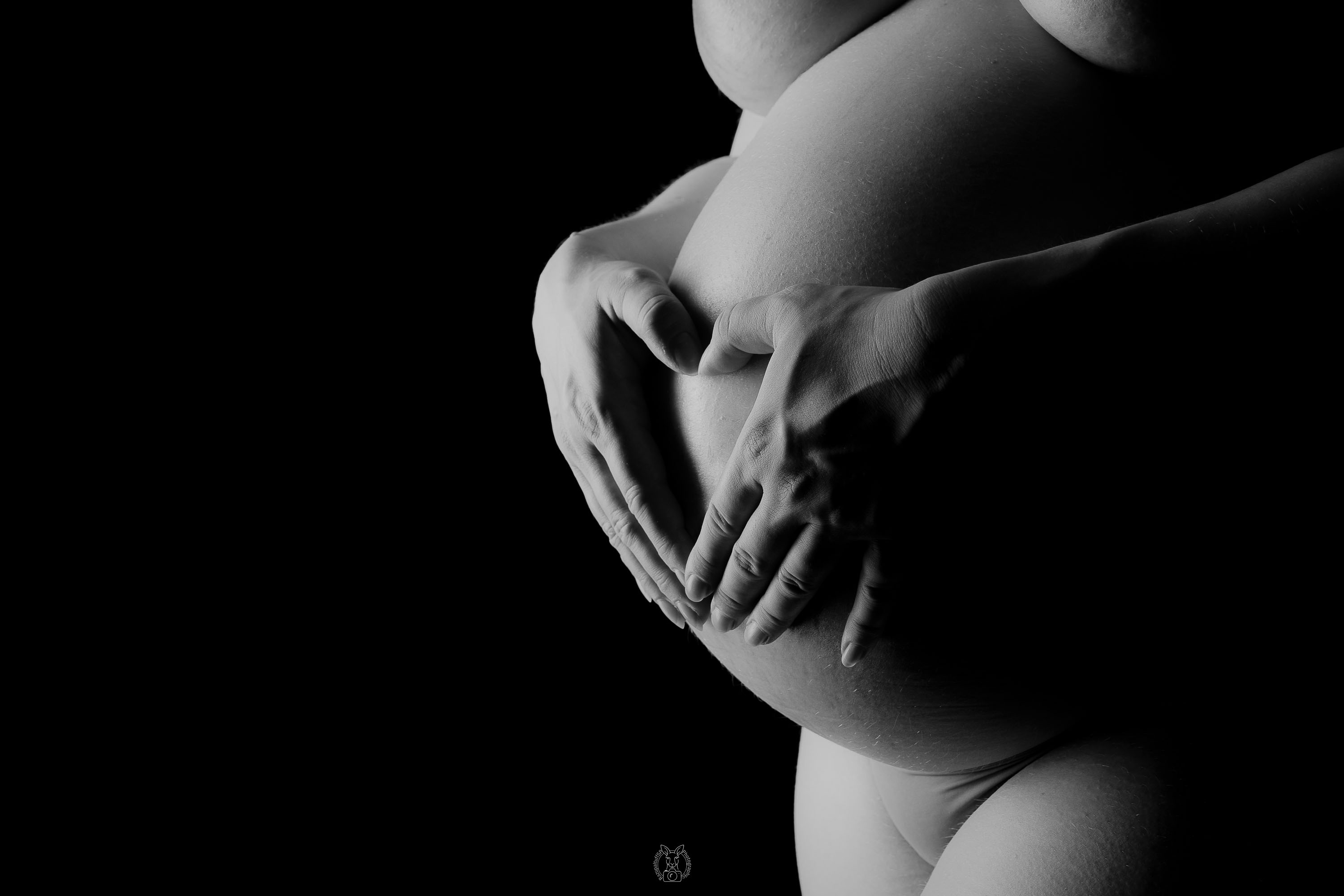 Nahaufnahme eines schwangeren Bauchs, umschlossen von zwei Händen, die ein Herz formen. Die Aufnahme ist in Schwarz-Weiß gehalten, mit starkem Kontrast.