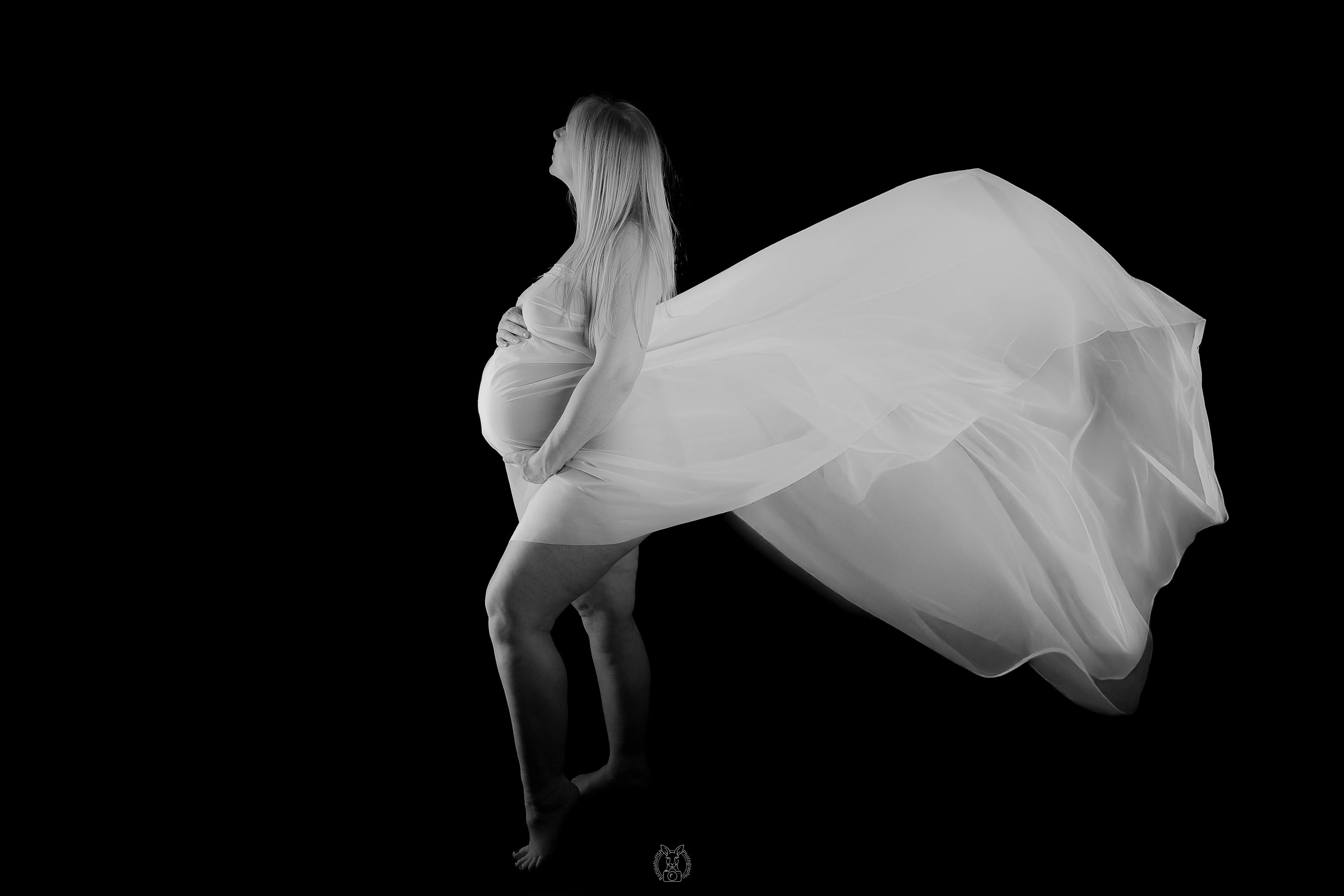 Eine schwangere Frau steht im Profil, eingehüllt in einen leichten, wehenden Stoff. Ihr Blick ist erhoben, der Hintergrund ist vollständig schwarz, das Licht fokussiert ihren Körper.
