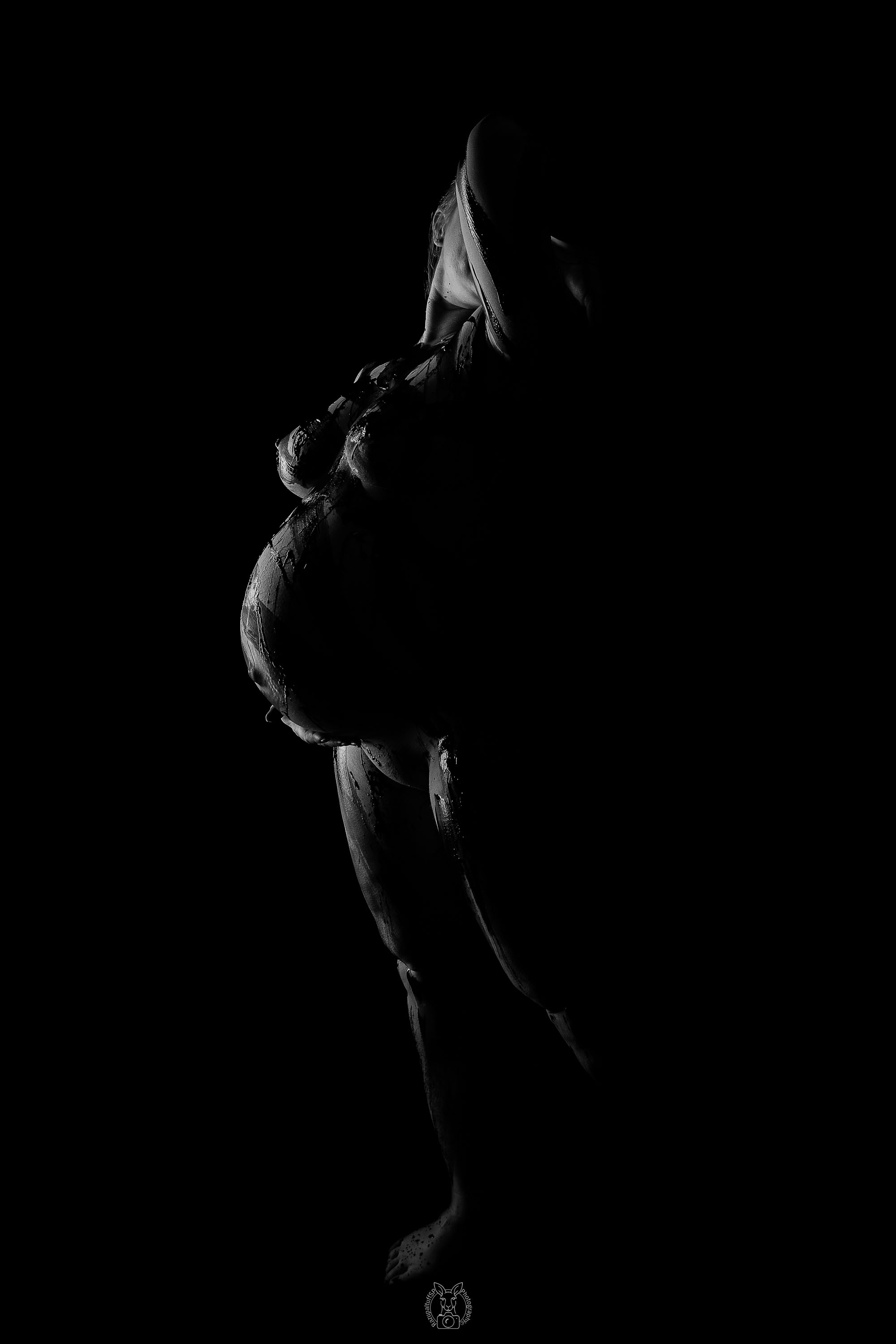 Dramatische Schwarz Weiß Aufnahme einer schwangeren Frau, deren Körper teilweise mit Farbe oder Ton bedeckt ist. Nur Teile des Körpers sind durch gezieltes Licht sichtbar.