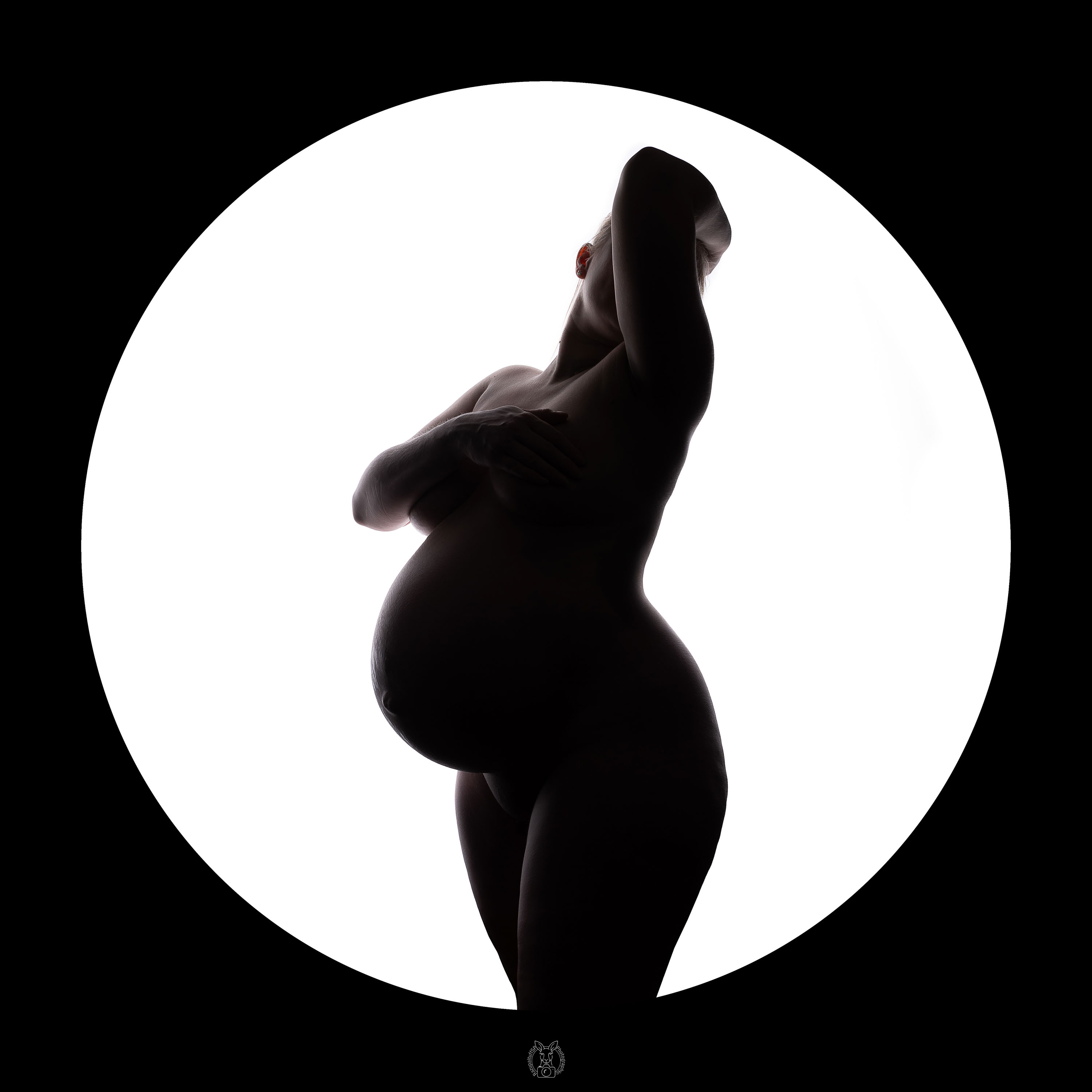 Eine schwangere Frau steht in einer runden Lichtfläche, der Rest des Bildes ist schwarz. Sie hält sich mit einem Arm über der Brust, der andere Arm ist erhoben. Ihr Babybauch ist stark betont.