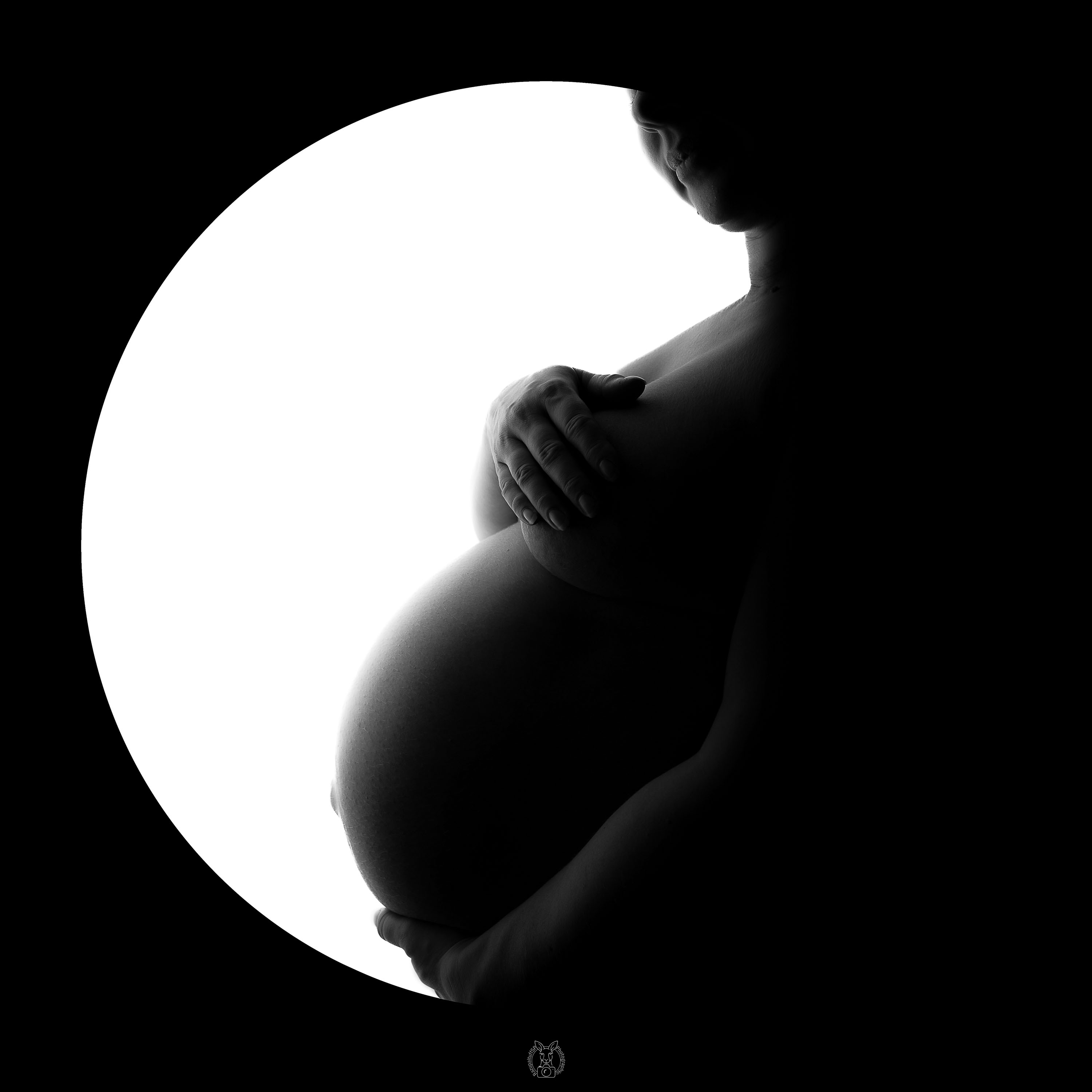 Schwangere Frau in Halbprofil, umgeben von einer runden weißen Lichtfläche, der Rest ist schwarz. Sie hält eine Hand auf der Brust und eine stützend unter den Bauch, mit einem sanften Lächeln im Gesicht.