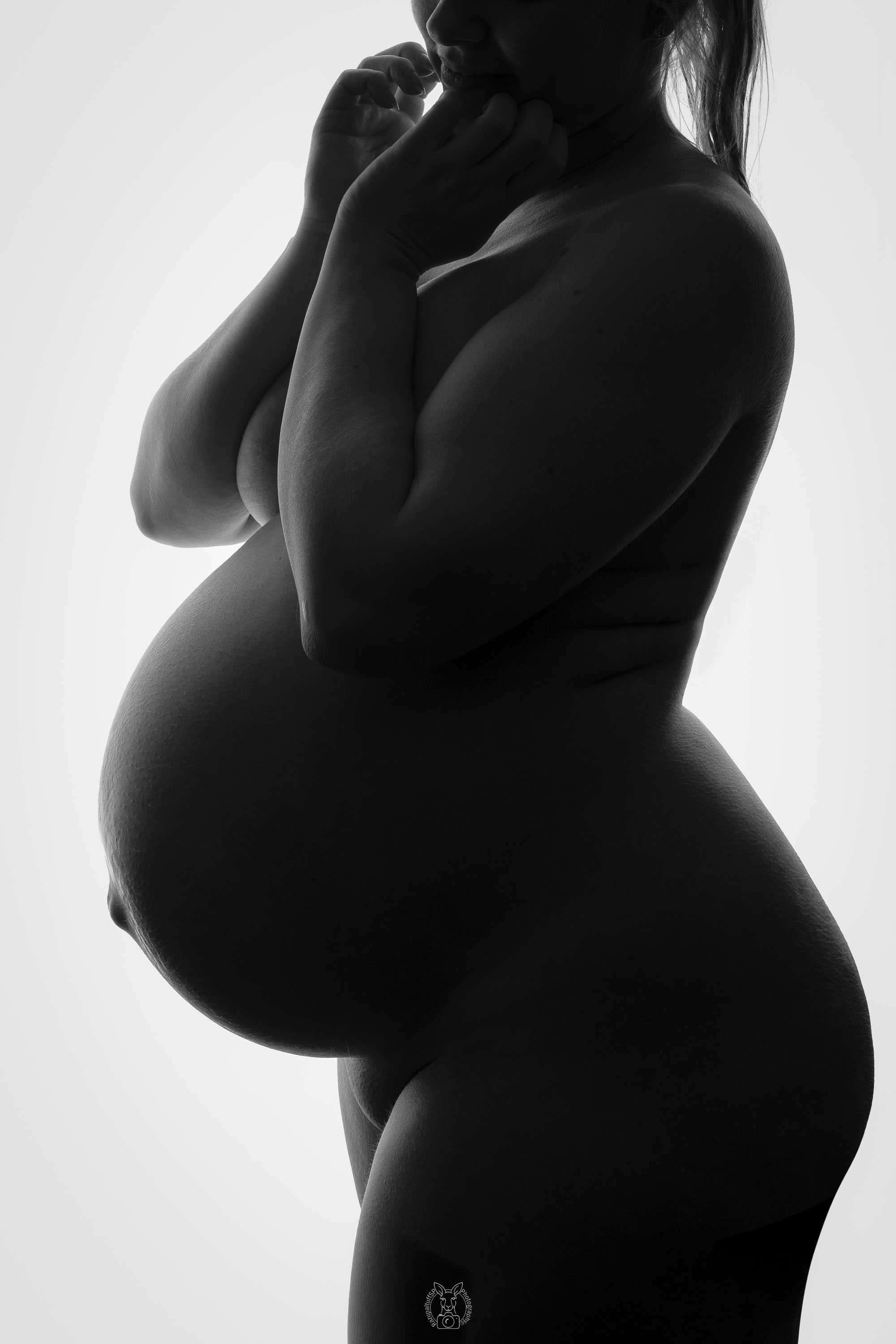 Silhouettenaufnahme einer schwangeren Frau in Seitenansicht. Sie hält beide Hände nah an ihr Gesicht, der Babybauch ist deutlich sichtbar. Die Lichtführung betont sanft die Körperformen.