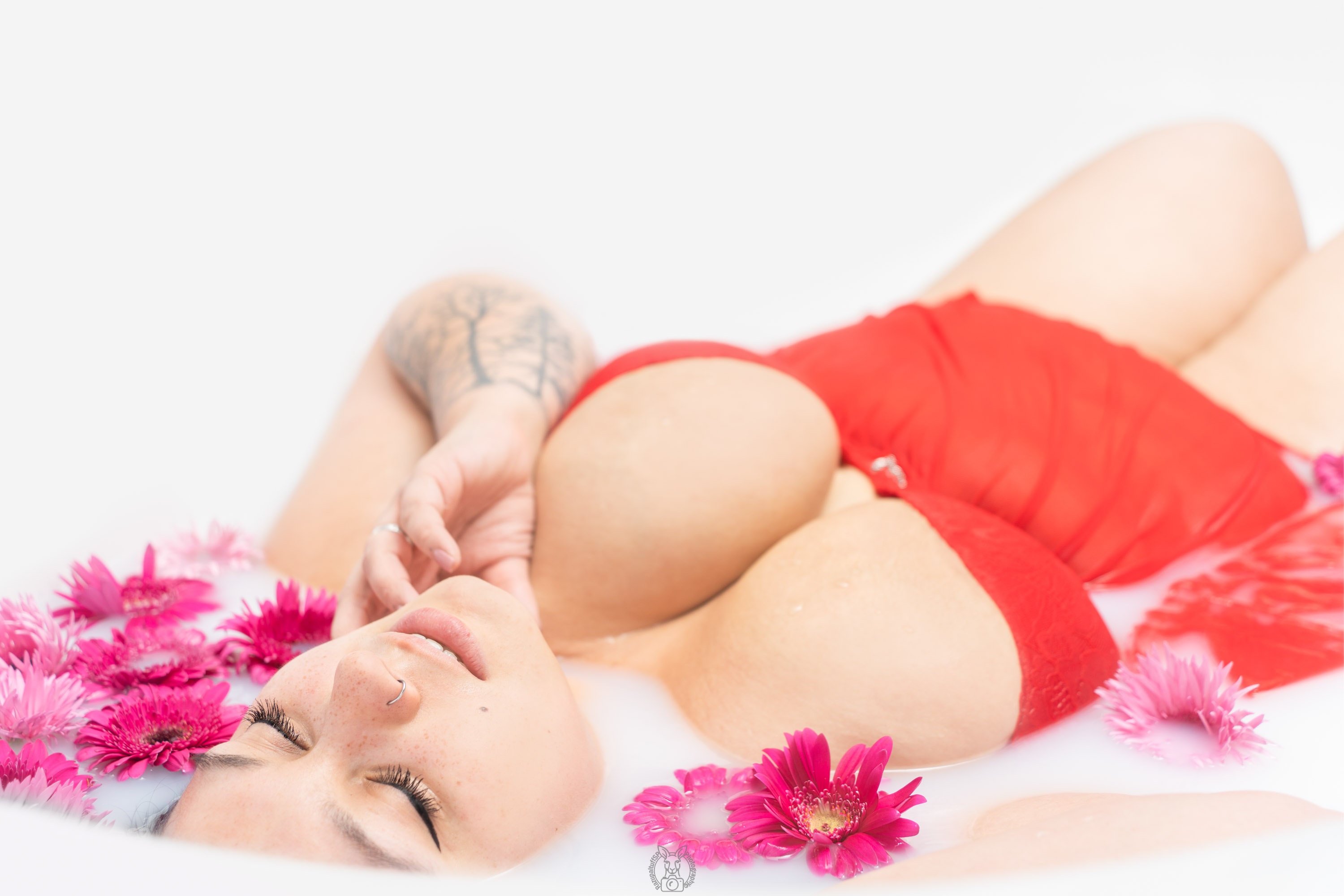 Milchiges Wasser umschließt den Körper. Rote Stoffe fließen weich über die Haut, umrahmt von kräftig pinkfarbenen Blumen. Eine Hand liegt am Gesicht, die Augen sind geschlossen – ein Moment innerer Ruhe.