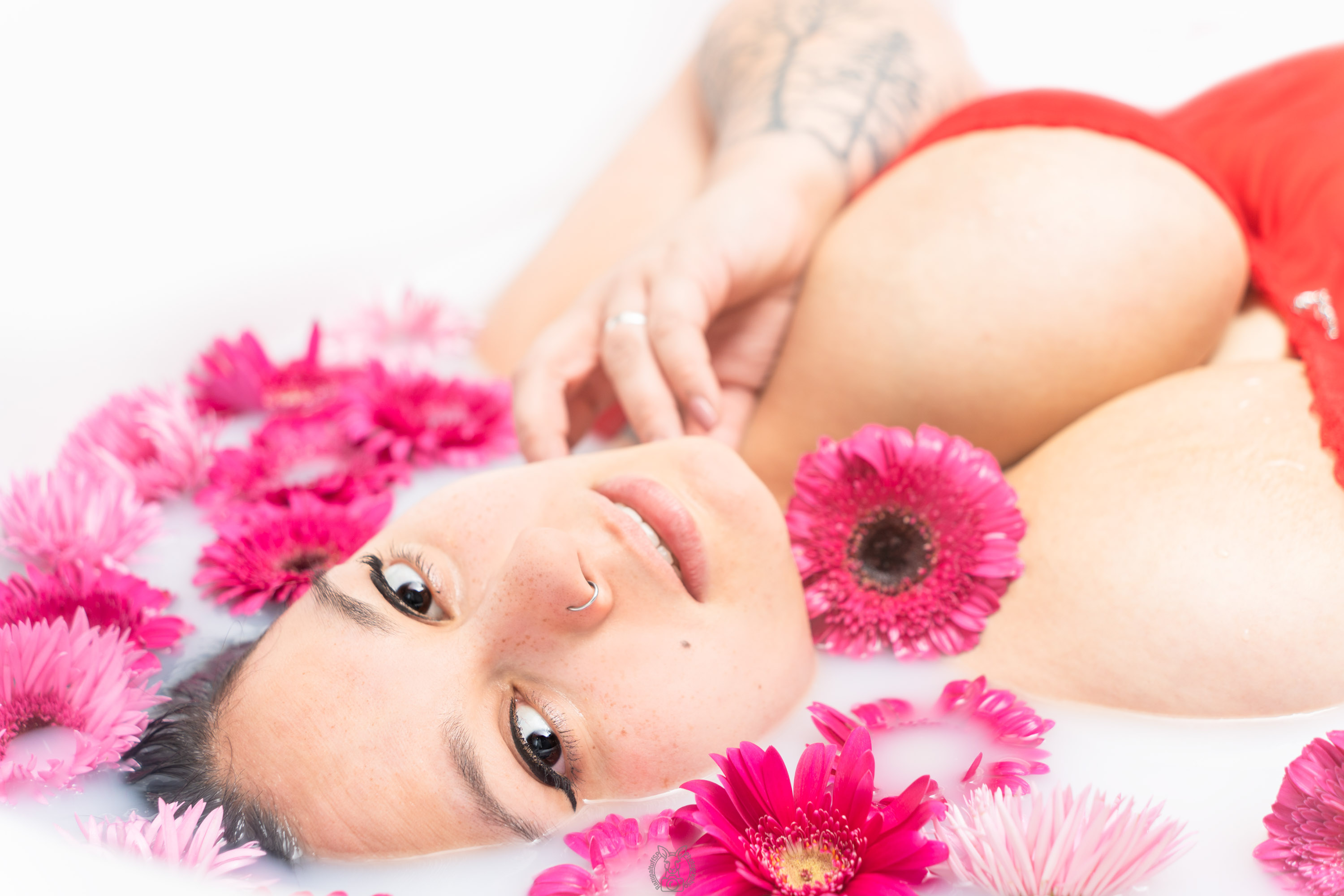 Der Blick ist wach und direkt. Der Kopf liegt leicht geneigt im Wasser, das Gesicht umgeben von leuchtenden Gerbera. Die Körperhaltung strahlt Sanftheit aus, das Licht betont die Konturen harmonisch.